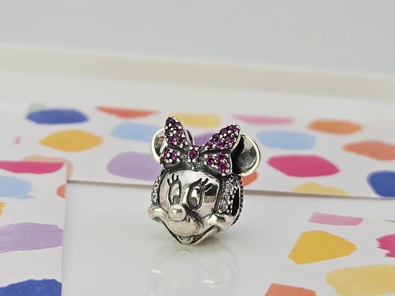 May include: A silver Minnie Mouse charm with a bow embellished with small, dark pink stones. The charm has a detailed design with small, clear stones around the face and ears. The charm is on a white surface with colourful shapes.
