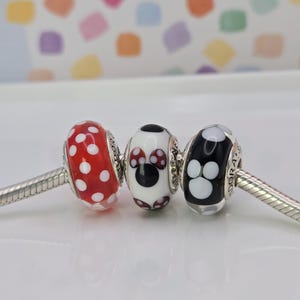 Puede incluir: Tres cuentas decorativas de vidrio en una cadena plateada. Una es roja con lunares blancos, la segunda es blanca con un dise&ntilde;o de Minnie Mouse y la tercera es negra con huellas de patas blancas.