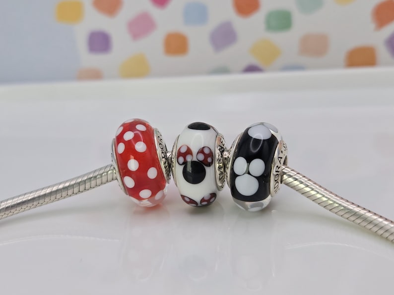 Puede incluir: Tres cuentas decorativas de vidrio en una cadena plateada. Una es roja con lunares blancos, la segunda es blanca con un dise&ntilde;o de Minnie Mouse y la tercera es negra con huellas de patas blancas.