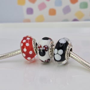 Puede incluir: Tres cuentas decorativas en una cadena de plata. Las cuentas son rojas con lunares blancos, una cuenta blanca con un dise&ntilde;o de Minnie Mouse y una cuenta negra con huellas de patas blancas. Las cuentas son para una pulsera.