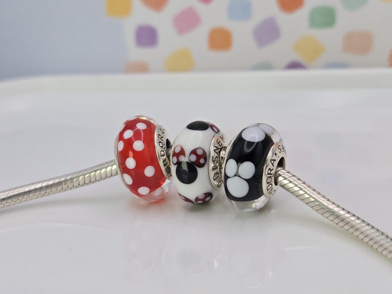 Puede incluir: Tres cuentas decorativas en una cadena de plata. Las cuentas son rojas con lunares blancos, una cuenta blanca con un dise&ntilde;o de Minnie Mouse y una cuenta negra con huellas de patas blancas. Las cuentas son para una pulsera.