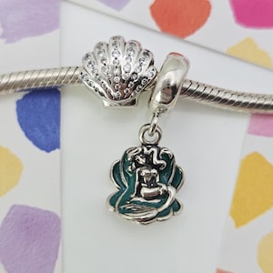Pode incluir: Pulseira de prata com três berloques. Um berloque é uma concha incrustada com pedras transparentes. Outro é uma conta de prata. O terceiro berloque é uma sereia com detalhes em verde-azulado.