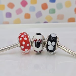 Puede incluir: Tres cuentas decorativas en una cadena plateada. Las cuentas son rojas con lunares blancos, un dise&ntilde;o de Minnie Mouse y una cuenta negra con huellas de patas blancas. El fondo es un patr&oacute;n colorido.