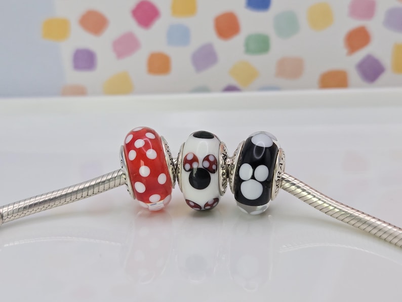 Puede incluir: Tres cuentas decorativas en una cadena plateada. Las cuentas son rojas con lunares blancos, un dise&ntilde;o de Minnie Mouse y una cuenta negra con huellas de patas blancas. El fondo es un patr&oacute;n colorido.