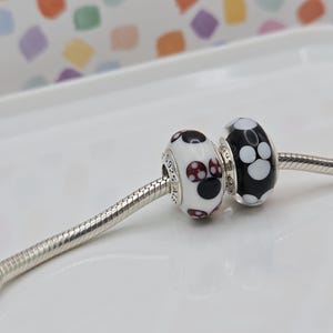 May include: Two decorative beads on a silver chain. One bead is white with black and burgundy polka dots. The other bead is black with white polka dots. The beads are likely for a bracelet or necklace.