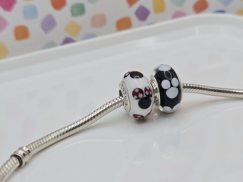 May include: Two decorative beads on a silver chain. One bead is white with black and burgundy polka dots. The other bead is black with white polka dots. The beads are likely for a bracelet or necklace.