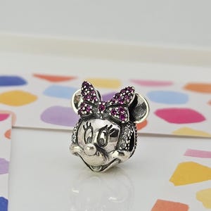 May include: A silver charm depicting Minnie Mouse. The charm features a bow adorned with purple gemstones and is detailed with small white gemstones. It has a polished finish and is set against a colourful background.