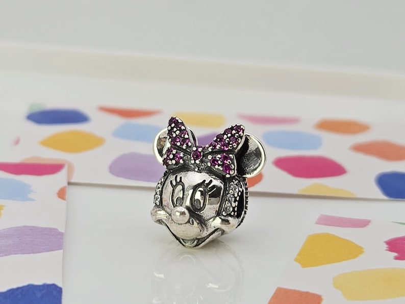May include: A silver charm depicting Minnie Mouse. The charm features a bow adorned with purple gemstones and is detailed with small white gemstones. It has a polished finish and is set against a colourful background.