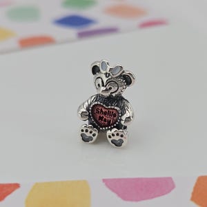 Könnte beinhalten: Ein silberner Charm-Anhänger in Form eines Bären mit einem Herz mit der Aufschrift "Shellie May". Der Bär hat eine Schleife und Pfotenabdrücke. Der Anhänger liegt auf einer weißen Oberfläche mit bunten Formen im Hintergrund.