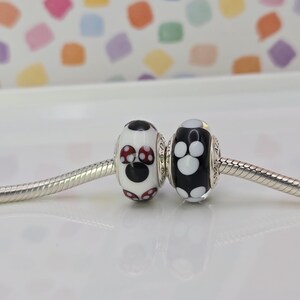 May include: Two glass beads with a whimsical design. One bead is white with a black circle and red polka dots, the other is black with white polka dots. Both beads are on a silver chain.