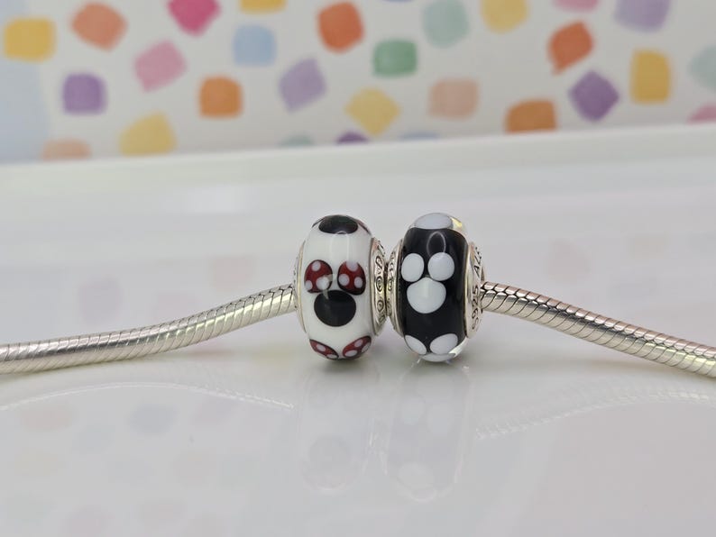 May include: Two glass beads with a whimsical design. One bead is white with a black circle and red polka dots, the other is black with white polka dots. Both beads are on a silver chain.