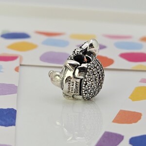 May include: A silver charm with a rounded, textured body encrusted with small, clear stones. The charm has a small, detailed head and a clasp. The charm is marked with "Pandora" and "Disney".