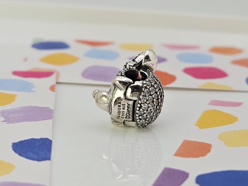 May include: A silver charm with a rounded, textured body encrusted with small, clear stones. The charm has a small, detailed head and a clasp. The charm is marked with "Pandora" and "Disney".