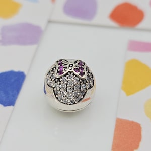 May include: A silver charm with a Minnie Mouse design. The charm features a bow with purple gems and a face made of clear, round gems. The charm is on a white surface with colorful watercolor splotches in the background.