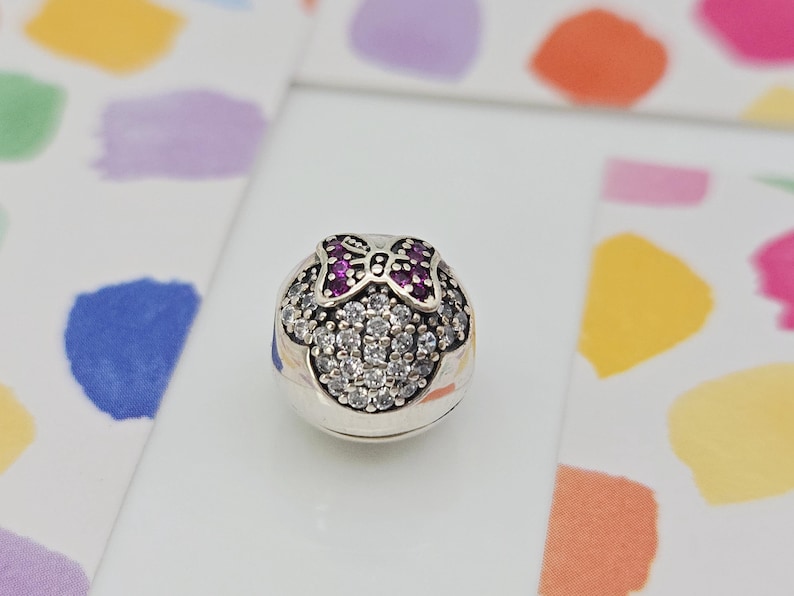 May include: A silver charm with a Minnie Mouse design. The charm features a bow with purple gems and a face made of clear, round gems. The charm is on a white surface with colorful watercolor splotches in the background.