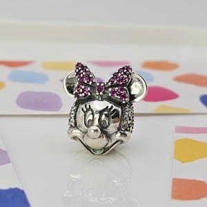 May include: A silver charm depicting Minnie Mouse's head. The bow is embellished with small, round, purple gemstones. The charm features a detailed face with large eyes and a button nose. The ears are round and smooth.