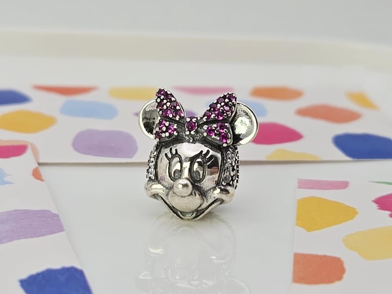 May include: A silver charm depicting Minnie Mouse's head. The bow is embellished with small, round, purple gemstones. The charm features a detailed face with large eyes and a button nose. The ears are round and smooth.