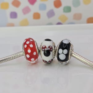 Puede incluir: Tres cuentas decorativas de vidrio en una pulsera plateada. Una cuenta es roja con lunares blancos, la segunda es blanca con un dise&ntilde;o de Minnie Mouse y la tercera es negra con huellas de patas blancas.