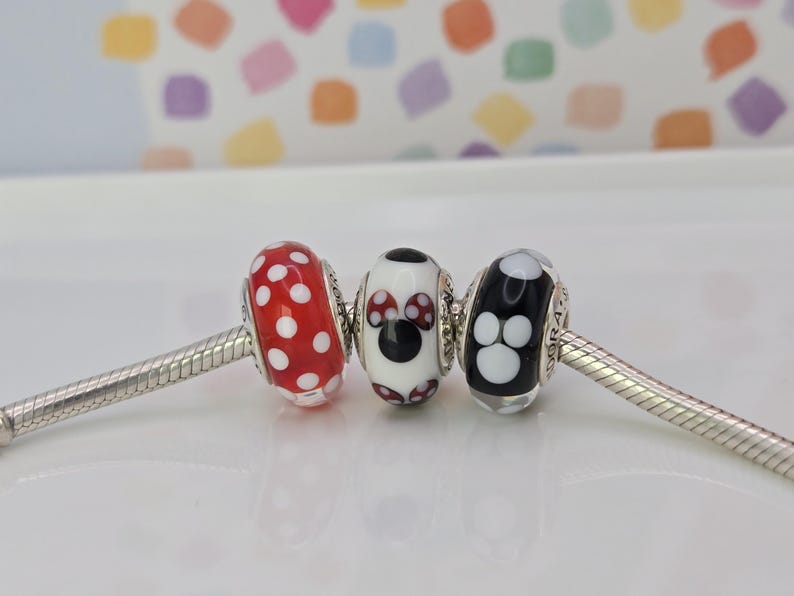 Puede incluir: Tres cuentas decorativas de vidrio en una pulsera plateada. Una cuenta es roja con lunares blancos, la segunda es blanca con un dise&ntilde;o de Minnie Mouse y la tercera es negra con huellas de patas blancas.