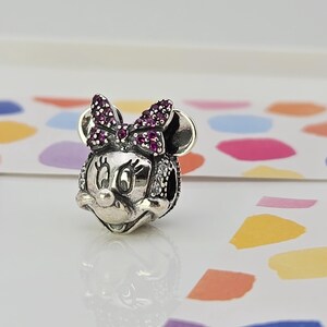 May include: A silver Minnie Mouse charm with a bow embellished with small, round, purple gemstones. The charm features a detailed face with black eyes and a small nose. The charm is on a white surface with colourful shapes in the background.