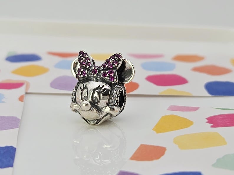 May include: A silver Minnie Mouse charm with a bow embellished with small, round, purple gemstones. The charm features a detailed face with black eyes and a small nose. The charm is on a white surface with colourful shapes in the background.