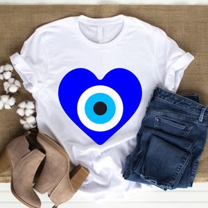 May include: White t-shirt featuring a large blue heart with an eye design. The shirt is paired with blue jeans and tan ankle boots. Cotton sprigs are arranged on a brown burlap surface.