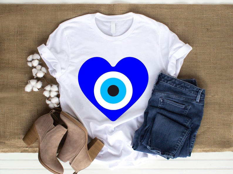 May include: White t-shirt featuring a large blue heart with an eye design. The shirt is paired with blue jeans and tan ankle boots. Cotton sprigs are arranged on a brown burlap surface.