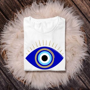 May include: White t-shirt featuring a large blue eye design with gold accents and radiating lines. The eye has a black pupil, surrounded by concentric circles of light blue and gold. The shirt is folded and rests on a fluffy white surface, set against a wooden background.