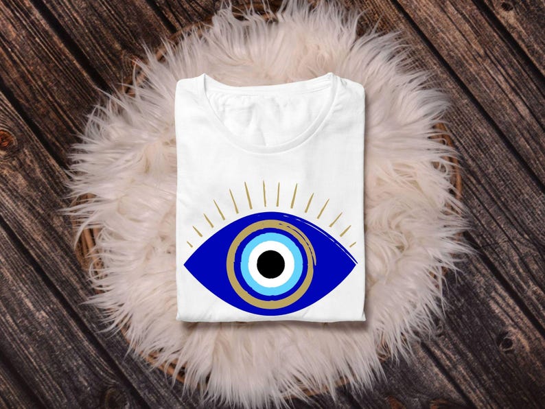 May include: White t-shirt featuring a large blue eye design with gold accents and radiating lines. The eye has a black pupil, surrounded by concentric circles of light blue and gold. The shirt is folded and rests on a fluffy white surface, set against a wooden background.