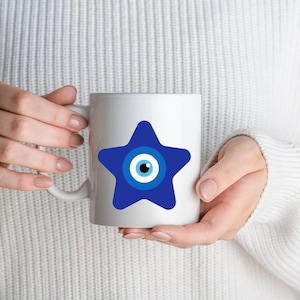 May include: White ceramic mug featuring a blue star design with an eye symbol in the center. The mug is held by a person wearing a white sweater. The mug is a standard 11 oz size.