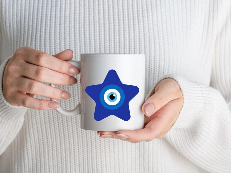 May include: White ceramic mug featuring a blue star design with an eye symbol in the center. The mug is held by a person wearing a white sweater. The mug is a standard 11 oz size.