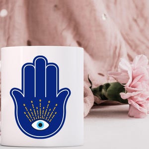 May include: White ceramic mug featuring a blue Hamsa hand design with an eye in the center. The Hamsa hand has a white outline and gold accents. A pink flower and pink fabric are in the background.