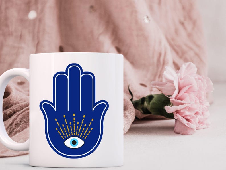 May include: White ceramic mug featuring a blue Hamsa hand design with an eye in the center. The Hamsa hand has a white outline and gold accents. A pink flower and pink fabric are in the background.