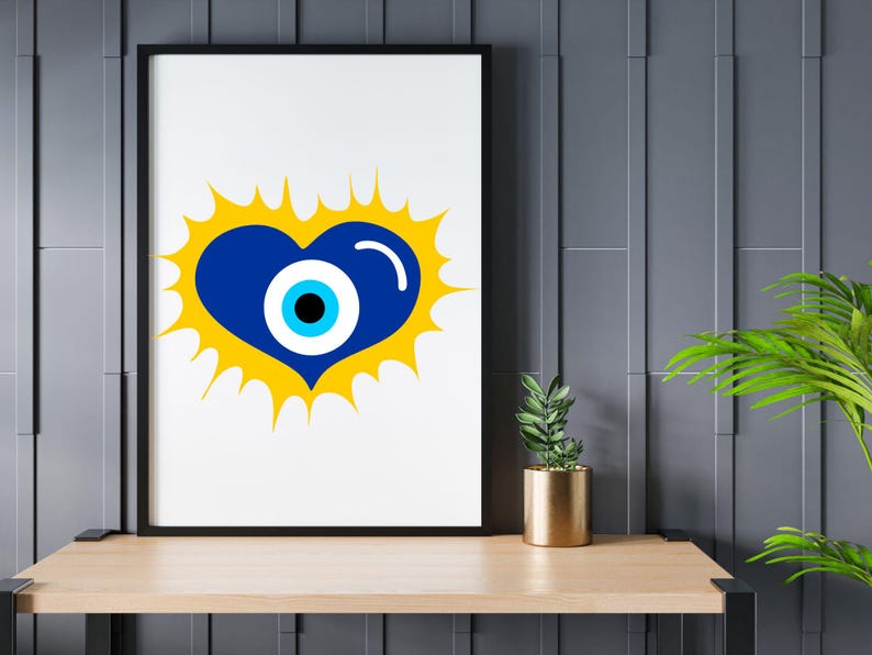 May include: A framed art print featuring a blue heart with an eye design, set against a yellow burst. The artwork is displayed on a light wood shelf with a small potted plant and a gold-colored pot. The background is a dark gray wall.