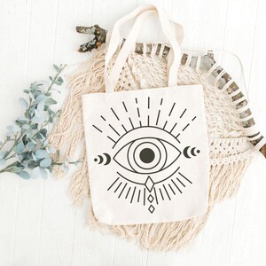 May include: Cream-colored tote bag with a black graphic of an eye with radiating lines, crescent moons, and a diamond shape. The bag has cream-colored handles. The bag is displayed on a white wooden surface with decorative elements.