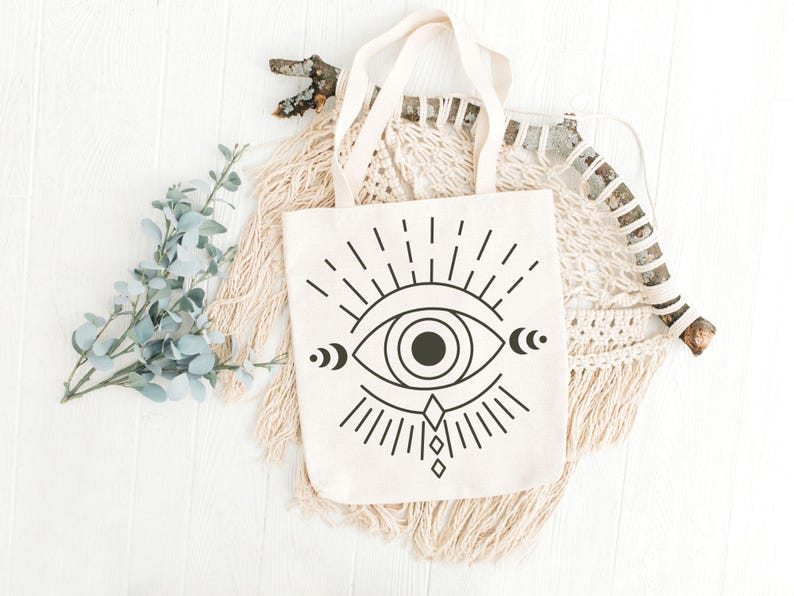 May include: Cream-colored tote bag with a black graphic of an eye with radiating lines, crescent moons, and a diamond shape. The bag has cream-colored handles. The bag is displayed on a white wooden surface with decorative elements.