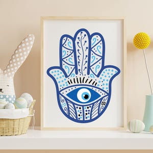 May include: A framed print featuring a blue Hamsa hand design with an eye in the center. The hand is decorated with intricate patterns and details. The artwork is displayed on a white surface, with a basket, decorative eggs, and flowers in the background.