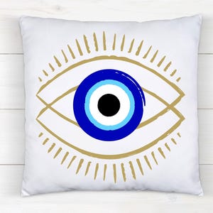 May include: White square pillow featuring a stylized eye design. The eye has a blue iris with a black pupil, surrounded by a gold-colored outline and radiating lines. The pillow is on a white wooden surface.
