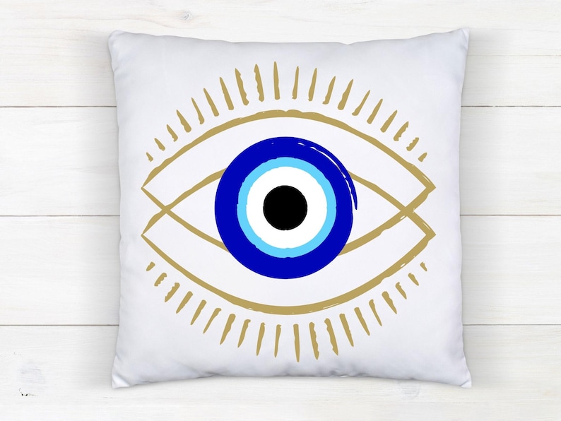 May include: White square pillow featuring a stylized eye design. The eye has a blue iris with a black pupil, surrounded by a gold-colored outline and radiating lines. The pillow is on a white wooden surface.