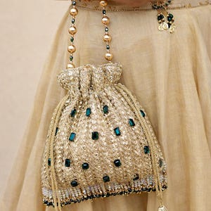 May include: Gold-colored drawstring bag with emerald green accents. The bag features a textured, embroidered design and is adorned with decorative tassels. The handle is made of gold beads and green accents. The bag is part of a formal outfit.