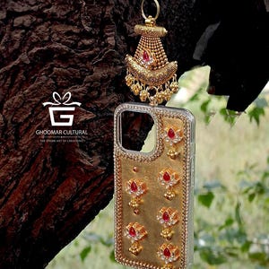May include: Ornate gold-coloured phone case with red and clear gemstones, and a matching charm. The case has a floral design and is embellished with small, round beads. The charm hangs from a gold-coloured clip.