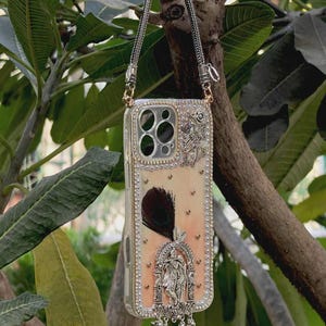 Puede incluir: Una funda decorativa para teléfono con una cadena plateada. La funda está adornada con pedrería, una pluma y una figura religiosa plateada. El fondo es de color melocotón claro.