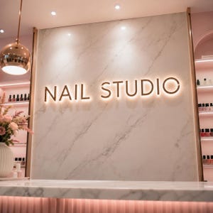 May include: A nail studio reception area featuring a white marble wall with backlit gold lettering that reads NAIL STUDIO. The space includes a white marble counter, a rose gold pendant light, and side shelves displaying various nail polish bottles.