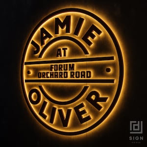 May include: A circular, illuminated sign with the text "JAMIE OLIVER" and "FORUM ORCHARD ROAD" in a dark brown font. The sign has a warm, golden glow, creating a striking contrast against the dark background. The sign also includes the text "SIGN VERSE".