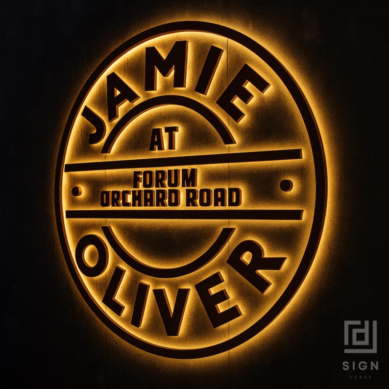 May include: A circular, illuminated sign with the text "JAMIE OLIVER" and "FORUM ORCHARD ROAD" in a dark brown font. The sign has a warm, golden glow, creating a striking contrast against the dark background. The sign also includes the text "SIGN VERSE".