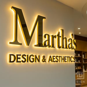 May include: A gold-colored illuminated sign on a wall. The sign reads "Martha's DESIGN & AESTHETICS" in a bold, sans-serif font. The letters are outlined with a warm, yellow glow, creating a striking visual effect.