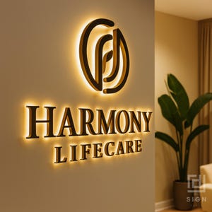 May include: Illuminated wall sign for "Harmony Lifecare" with a logo above the text. The sign is black with a warm, yellow glow emanating from behind the letters and logo. The sign is mounted on a light beige wall.