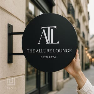 Puede incluir: Un letrero circular negro con texto blanco que dice "AL" encima de "THE ALLURE LOUNGE ESTD.2024". El letrero está montado en un soporte de metal negro en una pared beige. Una mano sostiene el letrero al aire libre.