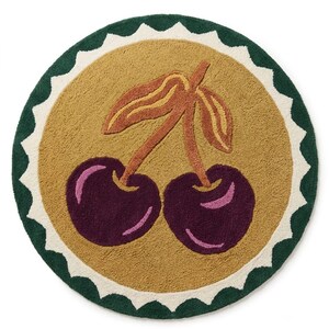 Puede incluir: Una alfombra circular con mechones que presenta un campo central de color amarillo mostaza con un gráfico de cerezas de color púrpura oscuro y dos tallos con hojas. La alfombra está bordeada por un borde festoneado blanco y un borde exterior verde.