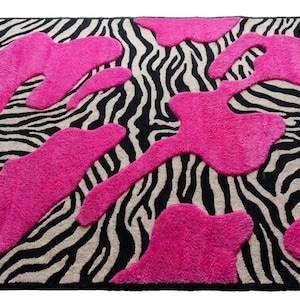 Puede incluir: Una alfombra rectangular con un patrón de rayas de cebra en negro y beige como base. Sobre la superficie se superponen varias formas irregulares, elevadas y con mechones de color rosa intenso que crean un efecto visual texturizado.